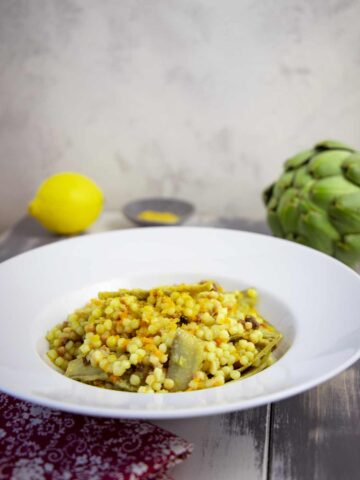 Teller mit Fregola-Pasta, Artischockenstücken und Bottarga auf dunklem Holztisch, im Hintergrund eine Zitrone und Artischocke.
