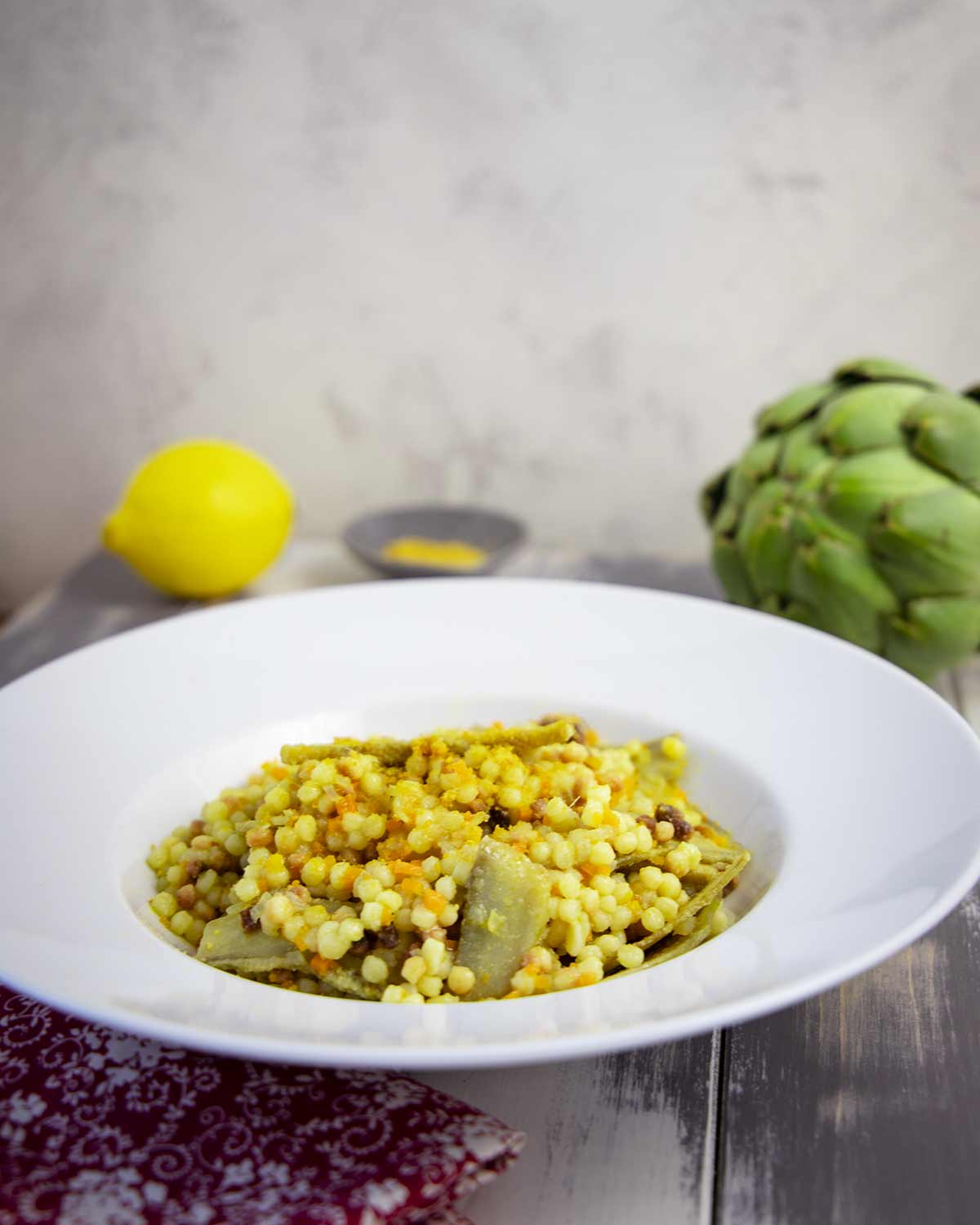Teller mit Fregola-Pasta, Artischockenstücken und Bottarga auf dunklem Holztisch, im Hintergrund eine Zitrone und Artischocke.