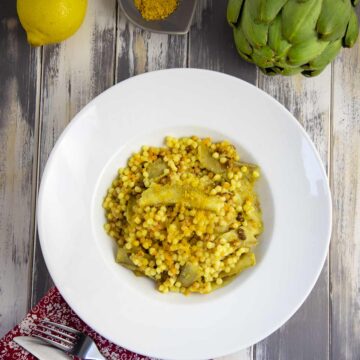 Teller mit sardischer Fregola mit Artischockenstücken, bestreut mit Bottarga. Daneben liegen eine Zitrone und Artischocke.